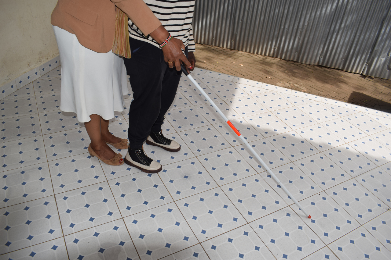 Photo of a blind man being helped to navigate the room with walking stick.